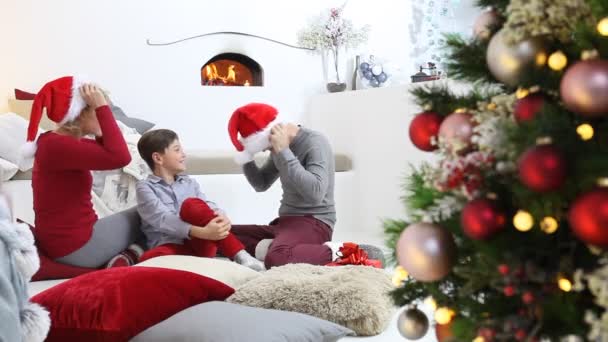 陽気なクリスマス 家で幸せな家族 両親はライトアップされた装飾された木の近くで息子と遊ぶ 喜びと笑顔がリビングルームに座って暖炉のそばで快適 — ストック動画