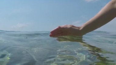 Ağır çekim yakın yarım sualtı çekim: su ile oynarken, parmakları ile yakalamak ve denize dökülen beyaz el. Dalgalar ve okyanus yüzeyinin üzerinde şaşırtıcı güneşli yaz gününde yansıtan güneş