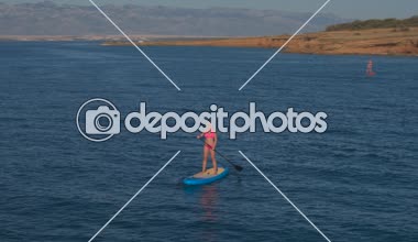 Hava: kadın paddleboarding çarpıcı kırmızı toprak cliffy duvar ve Doğal kayalık sahil boyunca yukarıda sıcak kuru yaz gününde uçan. Binici Sup sürme, raket tutarak ve eller havada kadar yükselen pembe bikini