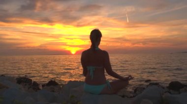 Yakın: kayanın üzerinde oturup deniz dalgalanan, lotus yoga poz meditasyon, konsantre, nefes ve rahatlatıcı zihin, ruh ve beden ateşli parlayan güneş ve altın ayarı gökyüzü altında gözlem yaparak sakin kız