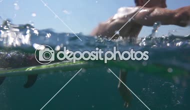 Ağır çekim yarım su altı: Açık deniz okyanus sörf tahtası üzerinde kürek çekmeye genç adam. Gülümseyen ve yaz tatillerini deniz kenarında üzerinde kristal berrak mavi denizi sanatçısını kürek çekmeye neşeli Erkek sörfçü