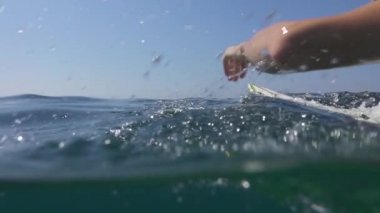 Ağır çekim yarım su altı: Açık deniz okyanus sörf tahtası üzerinde kürek çekmeye genç adam. Gülümseyen ve yaz tatillerini deniz kenarında üzerinde kristal berrak mavi denizi sanatçısını kürek çekmeye neşeli Erkek sörfçü