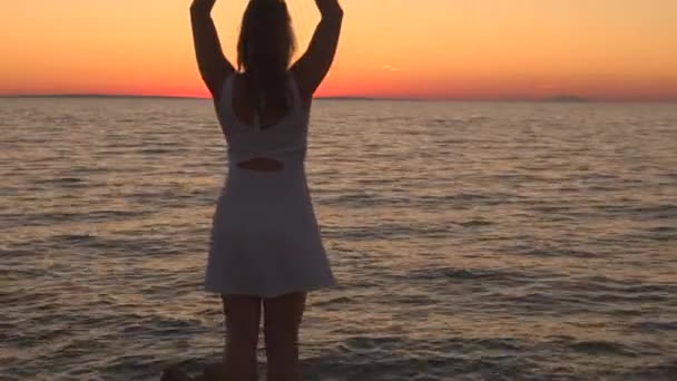白いドレス岩海海岸に立って 手でハートマークを作って 夕焼けで燃えるような金色の空を反映して見事な波打つ海を観察することで美しい女性の上を飛んで空中 クローズ アップ — ストック動画
