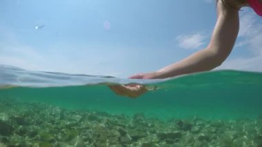 Ağır çekim yakın yarım sualtı çekim: su ile oynarken, parmakları ile yakalamak ve denize dökülen beyaz el. Dalgalar ve okyanus yüzeyinin üzerinde şaşırtıcı güneşli yaz gününde yansıtan güneş