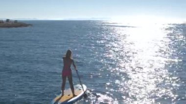 Hava, yakın: neşeli kız şişme standup paddleboard, sürme raket tutarak çevresinde ve eller havaya yükselen uçan. Deniz yüzeyi üzerinde şaşırtıcı güneşli yaz günü dalgalanan üzerinde yansıtan güneş