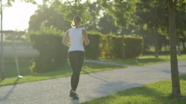 Ağır çekim Dof yakından: Koşu ve güzel park ağaçların arasında şaşırtıcı güneşli akşamı egzersiz şekil çekici genç kadın. Ağaç sağlayan canlandırıcı gölge sıcak yaz gününde Kanopiler
