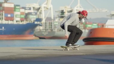 Ağır çekim yakın çekim: kaykay ve beton okyanus kıyısında güneşli günde arka seaport limanında demirlemiş yük gemisi gemi sürme heelflip hüner atlama Pro kaykaycı