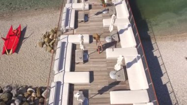Hava: Hafif yaz aylarında neşeli genç erkek giysi yürüyüş ve beyaz daybeds, tablolar ve şezlong ahşap iskele çarpıcı turkuaz deniz manzarası üzerinde boş lüks kokteyl barda arasında iplik