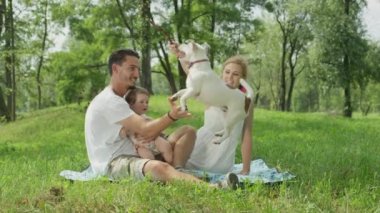 Ağır çekim, Close Up, Dof: Mutlu koyu saçlı Genç Baba, neşeli tatlı bebek kız ve battaniye park yere oturup, oyun ve sevimli köpek sopa ile alay beyaz elbiseli güzel sarışın anne
