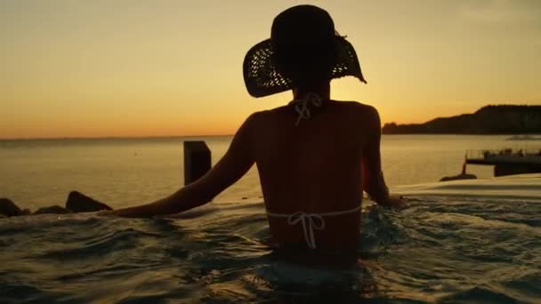 慢动作特写 有吸引力的金发碧眼的女人靠在无边泳池的边缘 欣赏金色的阳光落山 在高级海滨酒店的宁静夜晚 俯瞰迷人的岩石海洋海湾 — 图库视频影像