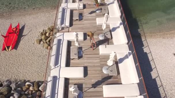 航空写真 光夏の陽気な若い女性ドレス歩くと白い寝台兼用の長椅子 テーブルと木製の桟橋が見事なターコイズ ブルーの海を見下ろす空の豪華カクテル バーのラウンジャー間回転 — ストック動画