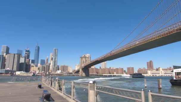 低角度のビュー ブルックリン橋の象徴的な表示やダウンタウンの高い現代的な高層ビル フェリーからマンハッタンをドッキングします ニューヨーク市のマンハッタンとブルックリンの自治区を接続するブルックリン橋 — ストック動画