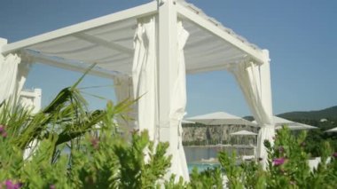 Ağır çekim yakın çekim: boş otel beach süslü Beyaz Köşk, daybeds, şezlong ve şemsiye pahalı lüks tatil güneşli yaz gününde çarpıcı. Mavi okyanus manzaralı sonsuzluk kenar havuzu