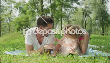 Ağır çekim yakın Up, Dof: Mutlu anne ve baba battaniye Park, çarpıcı güneşli bahar gününde pembe elbiseli neşeli tatlı bebek kız oynamaktan yatan portre. Çim bıçakla gıdıklıyor baba