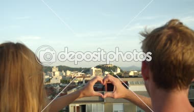 SLOW MOTION CLOSE UP DOF: Pretty girl with long hair and attractive boyfriend standing on edge of skyscraper rooftop kissing, holding hands and making heart shaped symbol with fingers at sunny evening