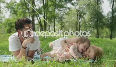 : Ağır çekim yakın kadar Dof mutlu genç baba ve güzel sarışın anne battaniye oynama ve pinky neşeli tatlı bebek kız ile gülüyor yerel parkta çim üzerinde yalan üzerinde güzel bahar günü giysi,