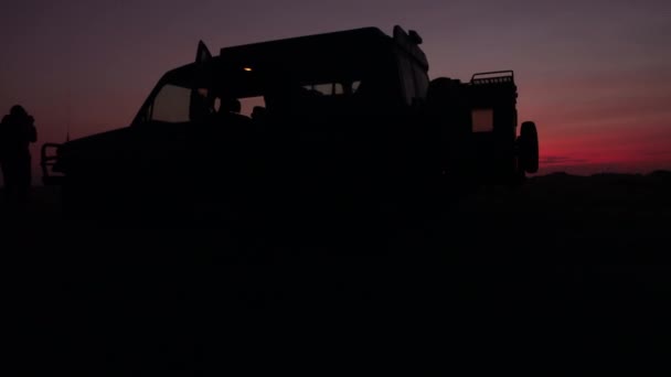 クローズアップ 夕暮れ時にアフリカの荒野の真ん中にオープン短い草サバンナフィールドに駐車2すべての地形サファリ車両のシルエット 魔法の黄金の光の夕日の写真を撮る観光客 — ストック動画