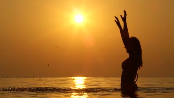 慢动作特写 欢快的女人在大海中站到腰部 在天空中举手 溅水滴 在神奇的金色夕阳下进行暑假活动 — 图库视频影像