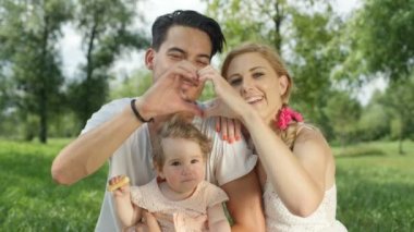 Ağır çekim Dof yakından: Oldukça neşeli anne ve sevimli gülümseyen Park, çimlerin üzerine oturan kalp elle yapma baba. Lezzetli kurabiye yemek kucağında oturan pembe elbiseli güzel mutlu bebek kız