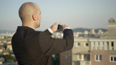 Ağır çekim yakın Up, Dof: binalar ve daireler satışa fotoğraflarını gökdelenin çatı üzerinde duran emlakçı. Anlık görüntüleri yapım onun smartphone üzerinde çalışan iş adamı