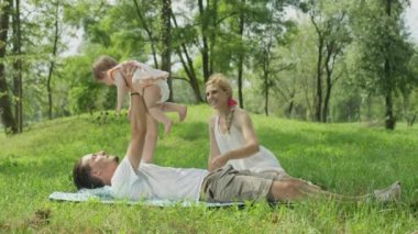 Ağır çekim yakın Up, Dof: oynayan neşeli tatlı bebek kız ile gür yeşil park battaniyeye yerde yatan genç baba. Gülümseyen ve kızının ayak gıdıklama yanında oturan beyaz elbiseli güzel annem