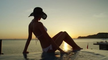 Ağır çekim yakın çekim: sonsuzluk havuzu kenarında oturan, su ile oyun ve altın güneş ayarı hayran çekici sarışın kadın. Yüksek sınıf oceanfront hotel İtalya huzurlu akşam