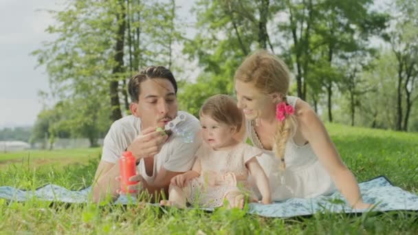 スローモーション クローズ アップ 被写し界深度 かわいい幸せな若い家族公園で遊んで芝生の上の毛布の上に横たわる 笑みを浮かべて美しい金髪の母親とかわいい女の子座っていると笑いを吹く陽気なお父さん — ストック動画