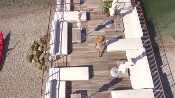 愉快的年轻女性穿着清淡的夏装 在白色的沙发床 桌子和躺椅之间散步和旋转 在木制码头上的空荡荡的豪华鸡尾酒吧里 可以俯瞰令人惊叹的绿松石海洋 — 图库视频影像