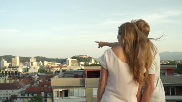 慢动作 一对夫妇站在屋顶的边缘 有很好的视野 看着美丽多彩的城市 男友带着他的家向女友指着手指向远处走去 — 图库视频影像