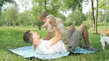 Ağır çekim, Close Up, Dof: yerel park battaniyeye arkasında yatan, neşeli tatlı bebek kız ile oynarken, onu havaya kaldırmış, onu yukarı ve aşağı atarak mutlu genç baba. Baba kız aile zaman