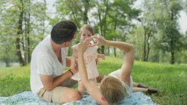 Ağır çekim Dof yakından: Genç Anne üzerinde park yapım kalp sembolü ile parmak, battaniyeye oturmuş ve çocuk tutan sempatik babası yalan. Pinky elbise gülümseyen içinde güzel neşeli kız bebek