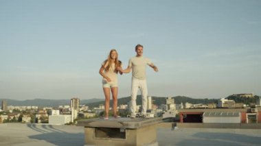 Ağır çekim yakın çekim: Pretty oynak kız ve boy gülümseyerek elele ve yüksek gökdelen çatı güneşli yaz sabahı çarpıcı üzerinde yeşil büyümüş tepelerle çevrili güzel şehir üzerinde atlama