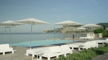 Ağır çekim, Close Up: boş otel beach süslü beyaz daybeds, şezlong ve şemsiye pahalı lüks tatil güneşli yaz gününde çarpıcı. Sonsuza kenar havuz bakan mavi sonsuz bir okyanus