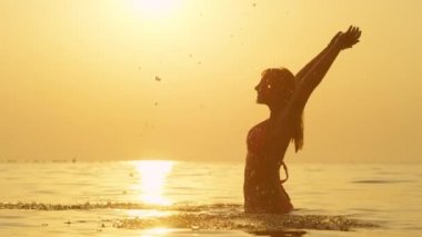 Slow Motion Close Up: Neşeli kadın denizde beline kadar ayakta, su kepçe, gökyüzünde ellerini yukarı kaldırarak ve su sıçraması etrafında damla. Büyülü altın gün batımında yaz tatillerinde aktiviteler
