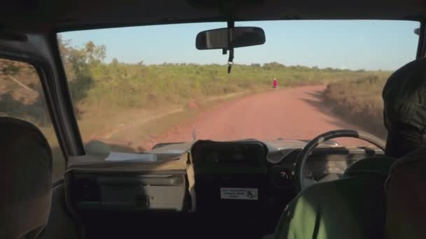 坦桑尼亚塞伦盖蒂 野生动物园吉普车驾驶游客在当地尘土飞扬的道路上驾车前往塞伦盖蒂国家公园 身穿传统红色衣服的马赛人沿路徒步 — 图库视频影像