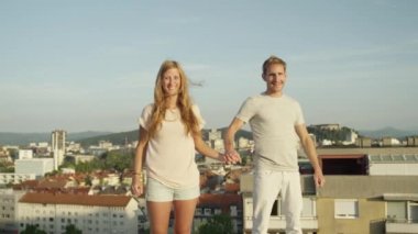 Ağır çekim kadar yakın: Heyecanlı kız ve elele ve onları üstünde yükselen güneş tarafından aydınlatılmış kentsel şehir üzerinde çatı yükseltmek boy gülümseyerek. Başarı kutluyor ve hayat sevgi dolu mutlu insanlar