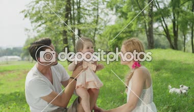 Ağır çekim, Close Up, Dof: onu mükemmel onun kollarında güneşli yaz günü holding neşeli bebek kız ile oynarken mutlu genç baba. Yanında oturan ve gülümseyen güzel neşeli anne. Sopa Çiğneme köpek