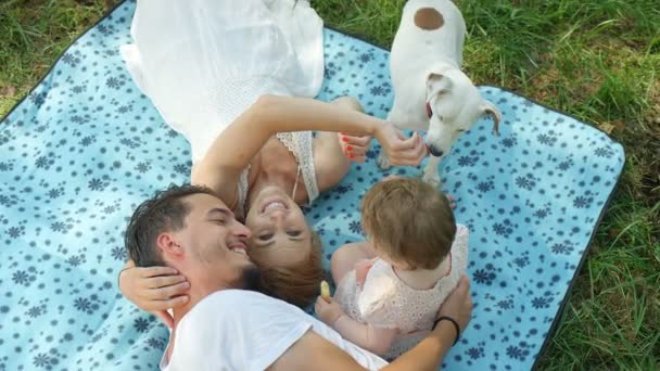 スローモーション クローズ アップ 愛らしい若い家族と晴れた日に遊ぶ庭に毛布の上に横たわってかわいい女の子 美しい母ふれあいの小型の犬は 幸せな父馴致娘の笑顔 — ストック動画