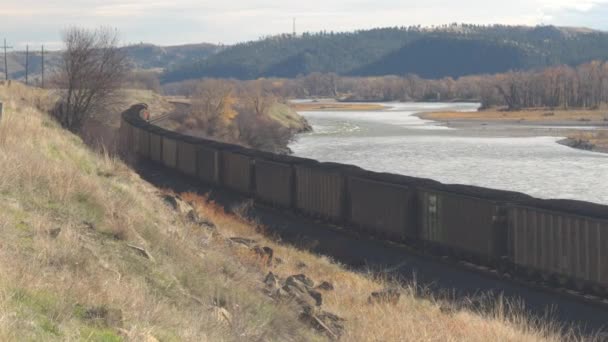 机车牵引一辆满载煤和焦炭的货运火车货车沿着风景如画的河流景观点缀着五颜六色的落叶树木和杂草丛生的山脉背景 — 图库视频影像