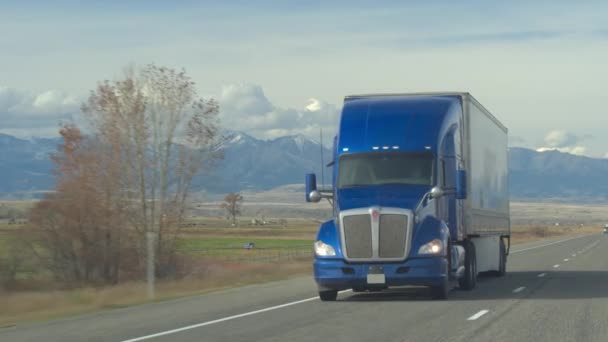 货运半卡车运输货物沿多车道公路穿过牧场和农田 在阳光明媚的日子里卡车卡车装载在背景岩石山脉范围内 — 图库视频影像