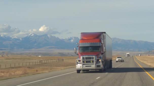 货运半卡车运输货物沿多车道公路穿过牧场和农田 在阳光明媚的日子 雪帽石山脉的背景下的卡车 — 图库视频影像