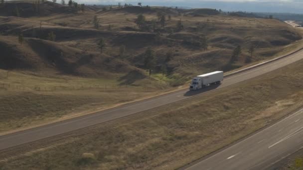 航空貨物コンテナー セミトラック日当たりの良いでこぼこ丘陵風景を介して実行している空のハイウェイに沿って運転の上を飛んでします アメリカ合衆国の高速道路の旅の間で配送コンテナーをトレーラーします — ストック動画