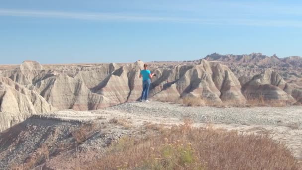 陽気な若い女性の旅行者は米国サウスダコタ州で晴れた日に風光明媚なバッドランズ国立公園の写真を撮影します 幸せな女の子砂岩山砂漠の荒野を散策やカメラで写真撮影 — ストック動画
