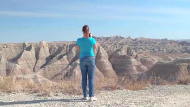 陽気な若い女性の旅行者は米国サウスダコタ州で晴れた日に風光明媚なバッドランズ国立公園の写真を撮影します 幸せな女の子砂岩山砂漠の荒野を散策やカメラで写真撮影 — ストック動画