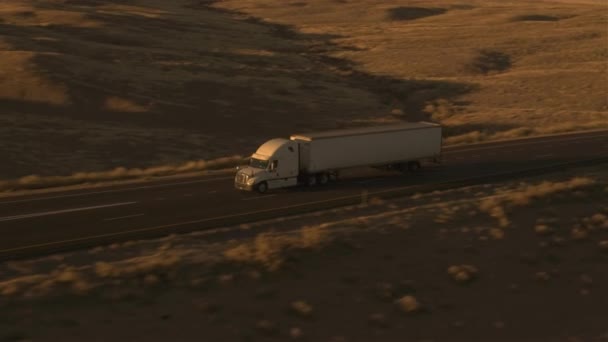 空中飞行在轮廓半拖车卡车与货运集装箱运载货物在公路驾驶通过平原草田航运货物横跨废弃的国家在黄金光黎明 — 图库视频影像