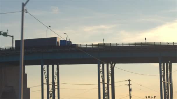 低角度看半卡车驾驶过天桥 运输货物横跨繁忙的多车道公路在金色日出 满载货车在蓝天上搬运货柜 — 图库视频影像