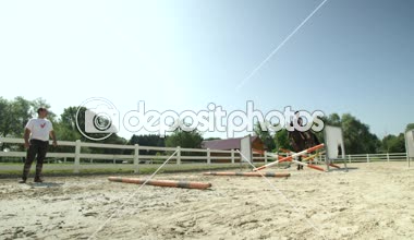 Ağır çekim yakın çekim: gözlemleyerek ve Genç horsegirl öğrenci birkaç art arda çit ve kumlu açık alanda engellerin üzerinden atlama zaman düzeltme eğitmeni. At atlama için kadın eğitim