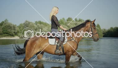 Ağır çekim Dof yakından: Uzun saç Binicilik at geniş Nehri güneşli yaz gününde çarpıcı güzel kızla. Genç kadın binici etrafında su sıçramasına kahverengi iğdiş edilmiş hayvan geçiş Nehri üzerinde