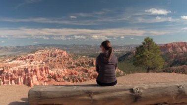 Gülümseyen kadın gezgin çarpıcı Bryce Canyon Amfitiyatro bakan ahşap bankta oturan. Bryce Canyon Milli Parkı'nda kumtaşı hoodoos ve kayalık sütunlar bakan pitoresk bakış açısı.