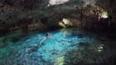 Tavandaki delikten gelen güneş ışığıyla sihirli yeraltı mağarasında turkuaz havuzda yüzen genç bir kadın. Aktun Chen 'deki sarkıtların ve dikitlerin büyüleyici mimarisi.